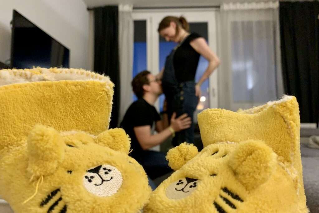 Im Hintergrund stehen die schwangere Autorin, Lasse kniet davor und küssten den dicken Bauch. Im Vordergrund zwei Babyschuhe in Tigerform.